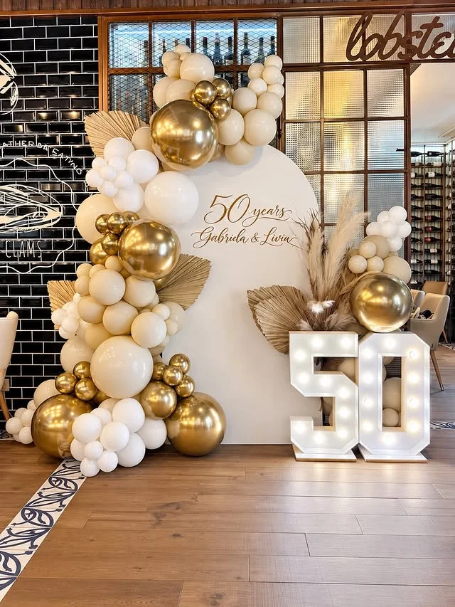 White and gold balloon arch with dried palms and light up 50 numbers at a golden celebration