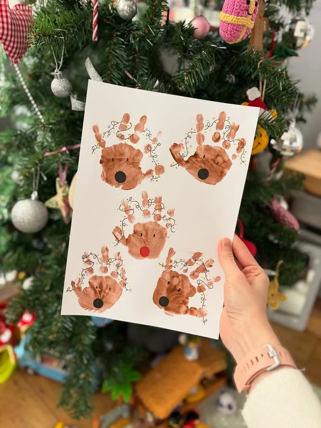 Handprint reindeer faces art with Christmas light antlers held in front of tree