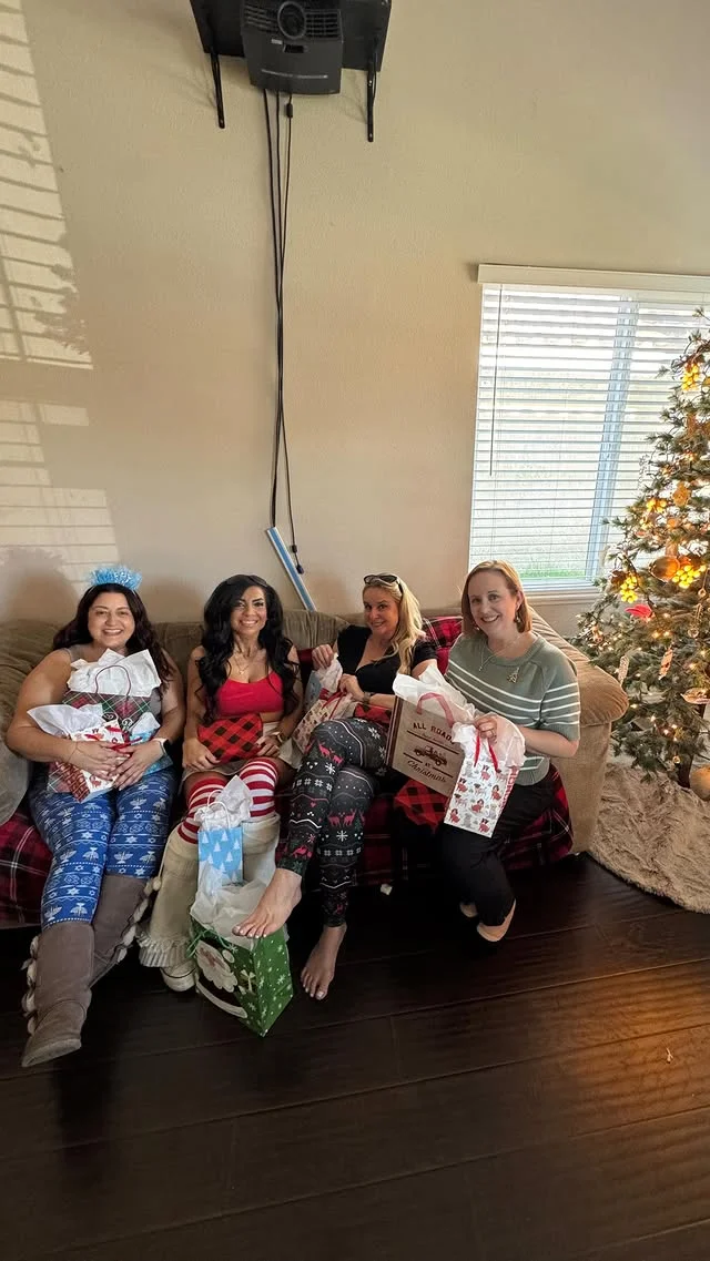 Four women on a couch each holding wrapped Christmas gifts beside a lit tree