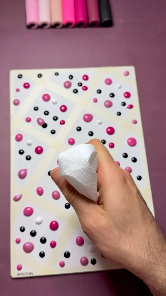Hand holding a paper towel over a tape grid canvas dotted with pink, black and white paint