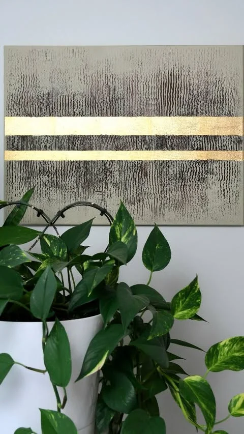 Textured gray abstract canvas with two horizontal gold leaf stripes hanging above a green plant