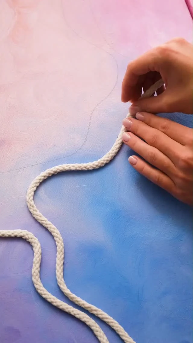 Hands laying a white rope in a curve over a pink and blue painted canvas