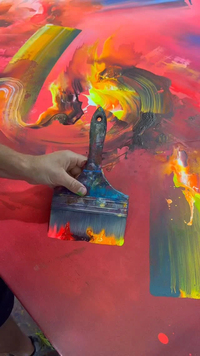 Hand holding a brush over a vivid abstract painting swept with red, yellow, blue and green paint
