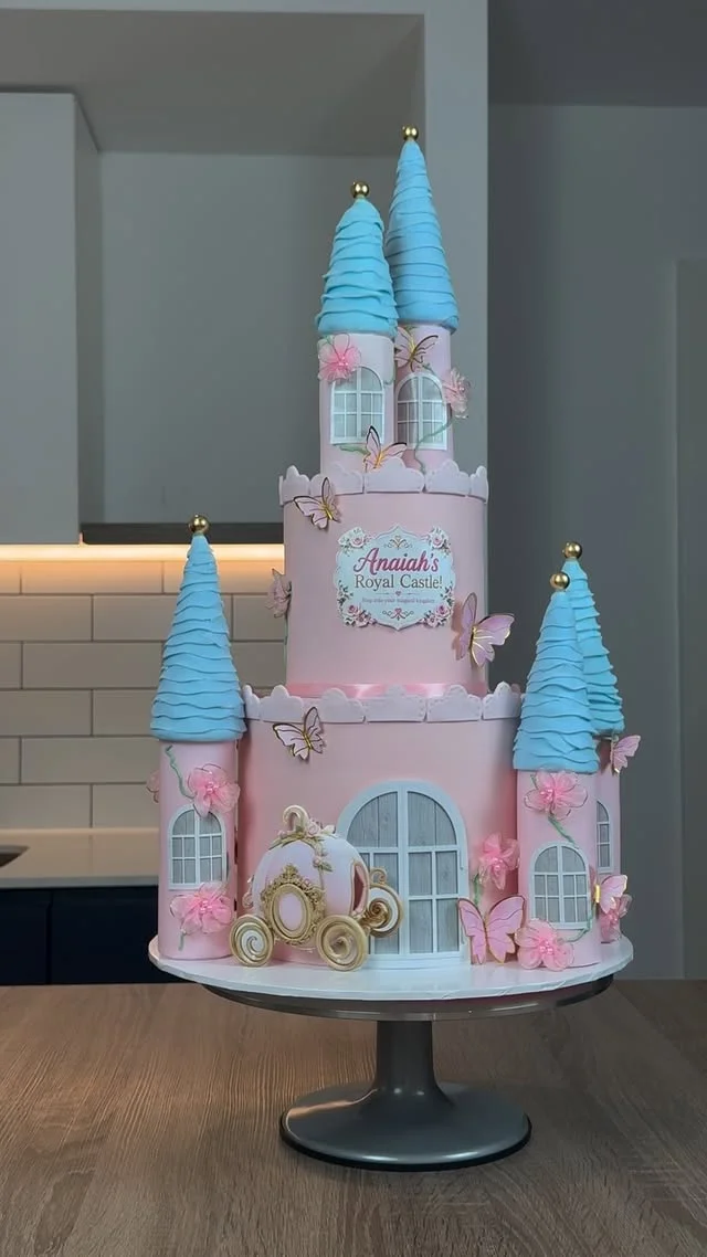 Pink castle cake with blue cone turrets, butterflies and a gold carriage on a cake stand