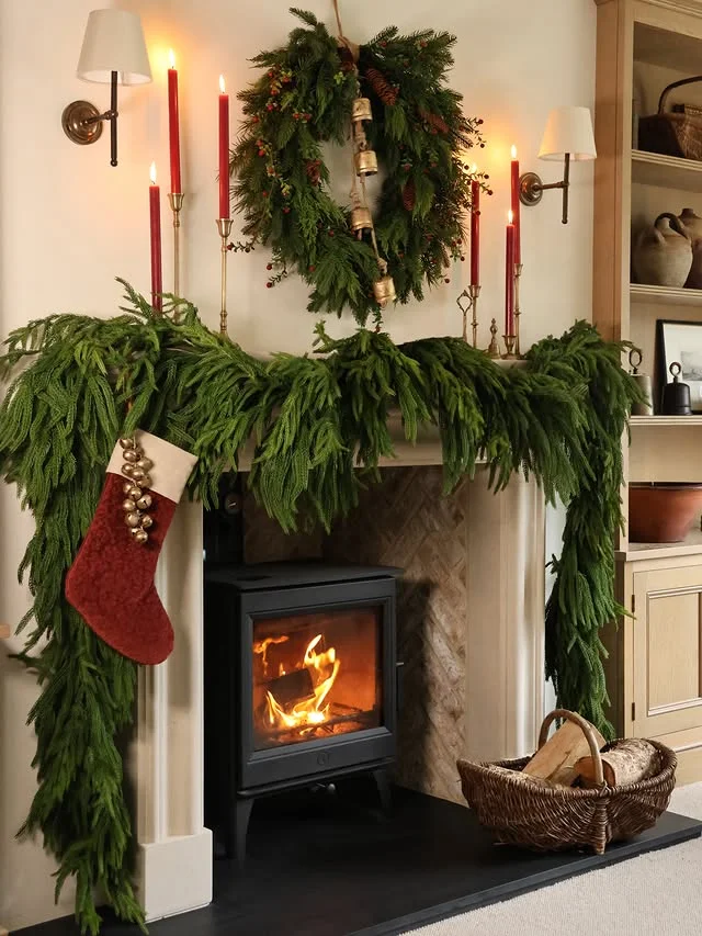 Elegant Christmas mantel styled with lush green garland, red taper candles in brass holders, a wreath with pinecones and brass bells, and a red velvet stocking