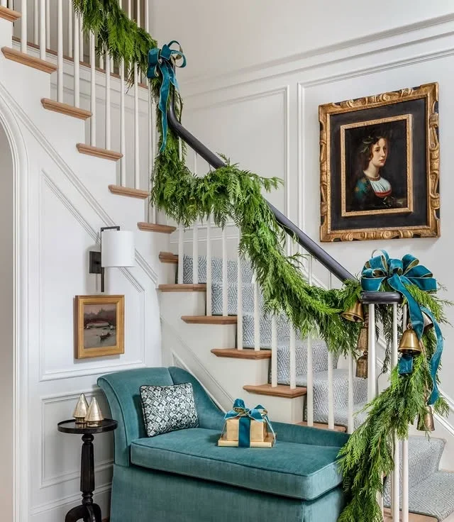 Elegant Christmas entryway with a white staircase draped in evergreen garland, teal velvet bows, gold bells, and a teal settee with a wrapped gift