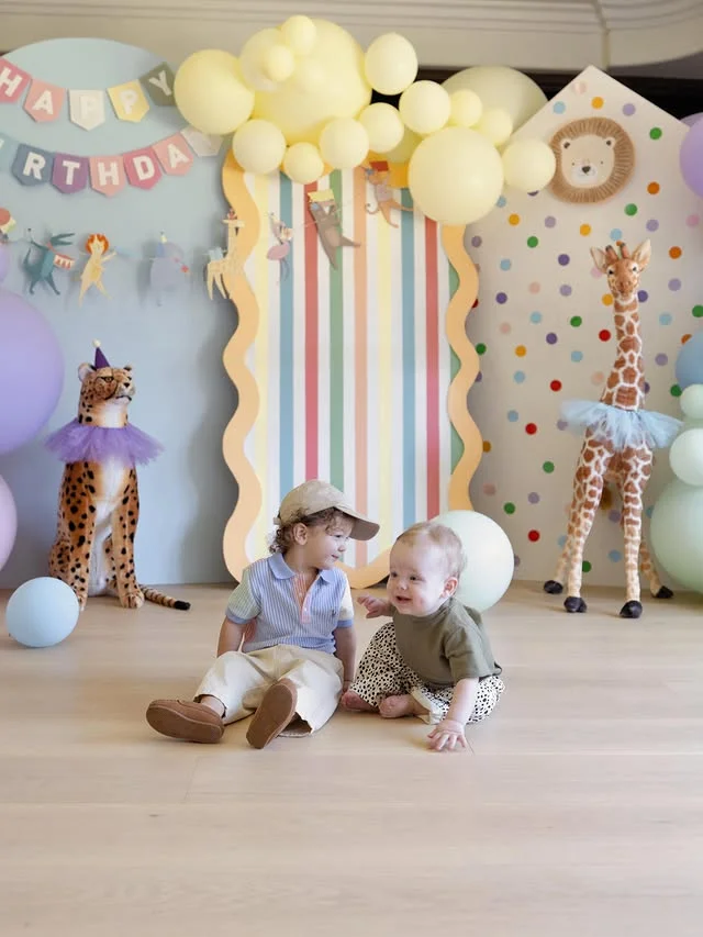 Pastel party animal backdrop with giraffe and cheetah figures and two toddlers